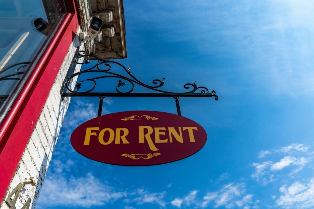 A view from below of a hanging red sign that says For Rent in fancy lettering and hangs from a white building, against the backdrop of a bright blue sky with some clouds.