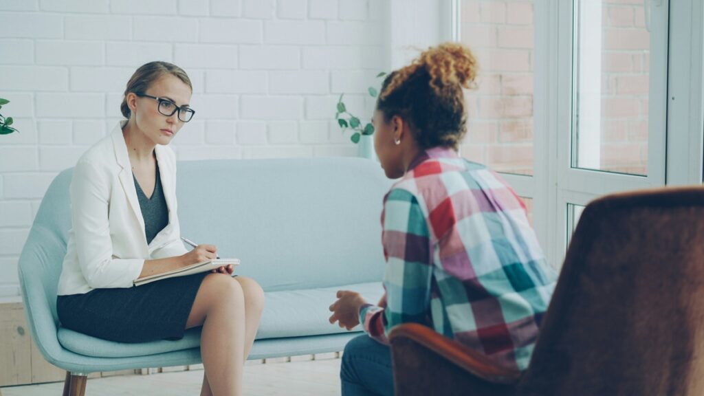 A female therapist sitting on a blue couch, with her hair in a bun and wearing glasses, takes notes as she talks to another woman, sitting on a brown chair.