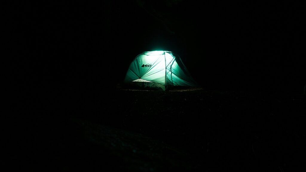 A green tent that says "REI" is lit from inside and sits against a dark sky.