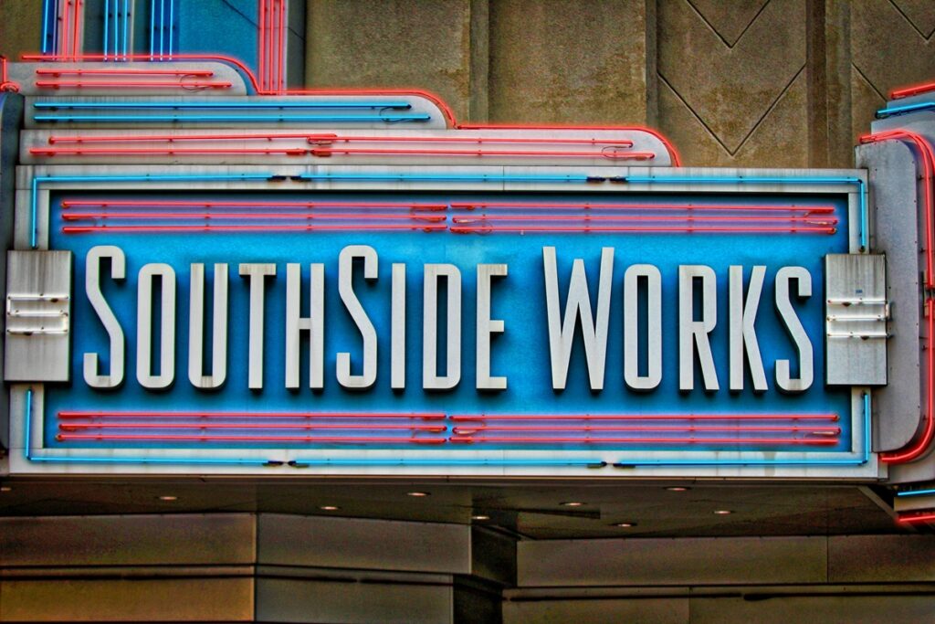 A photo of a marquee sign that says South Side Works. The sign is metal. South Side Works is white while the background is blue with red lines.