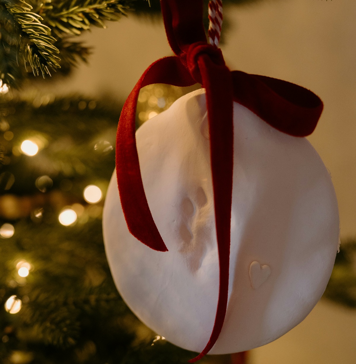 A close up of a clay ornament with a pet's pawprint. It's hanging in front of a Christmas tree with white lights