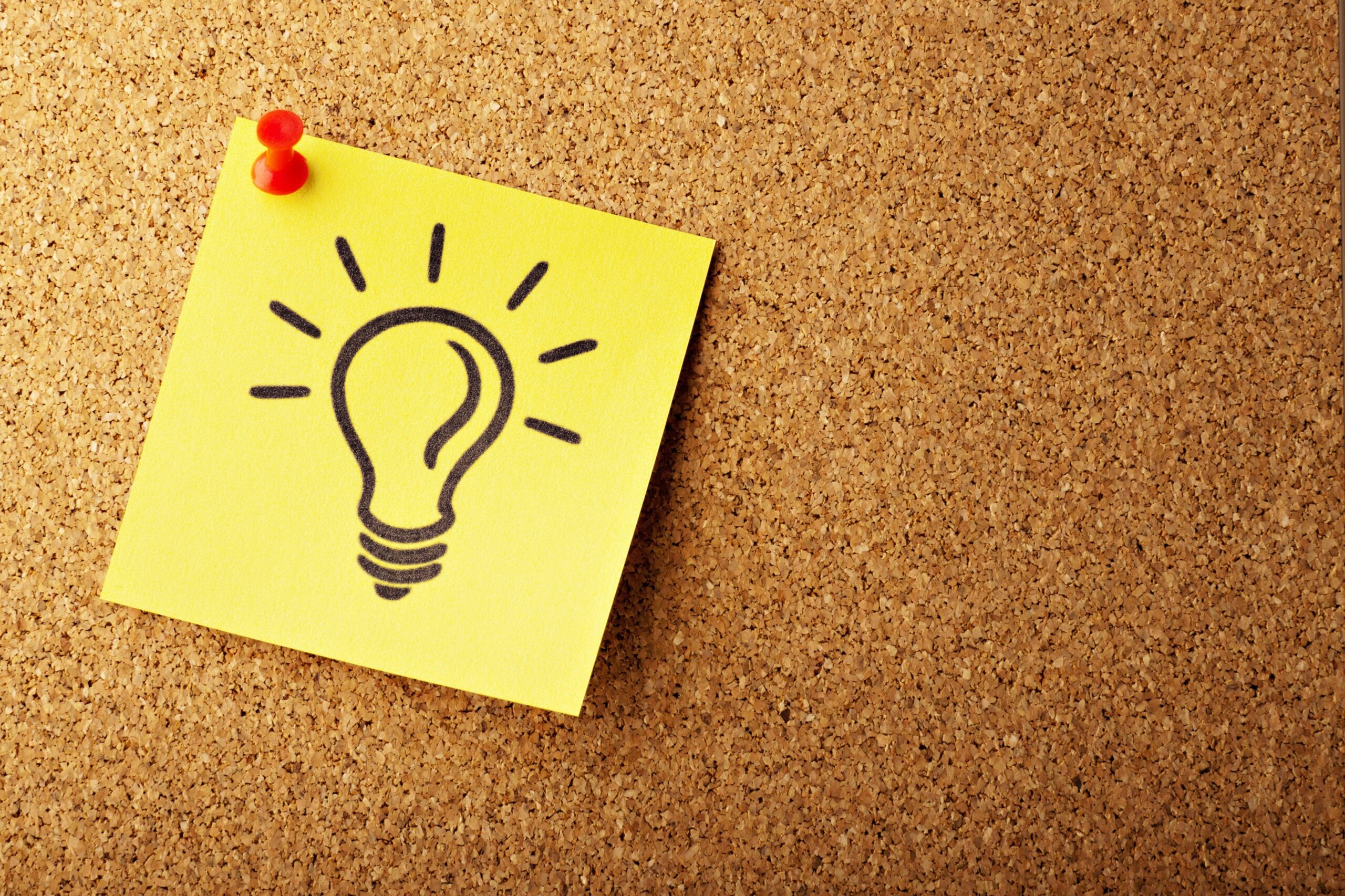 A photo of a cork board with a yellow sticky. The yellow sticky has a light bulb drawn on it and it's attached to the board by a red tack.