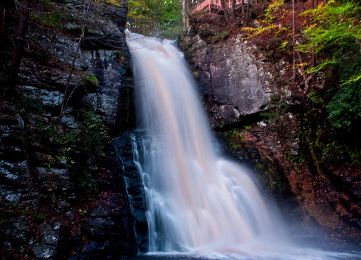 A photo of a waterfall dropping between two large slabs or rock. Above the rock on the right side is a wooden railing (part of a viewing platform).