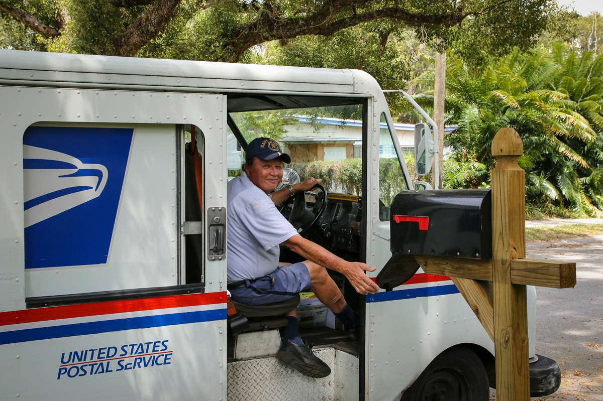 A mail carrier sits in his USPS mail truck and looks at the camera.