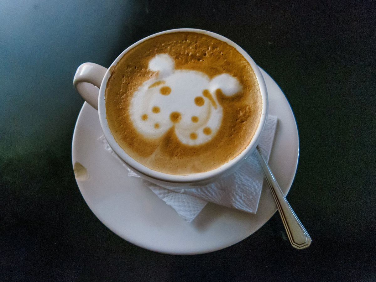 A cup of coffee on a white plate. The cup is sitting on two white napkins with a spoon to the right of the napkins. The face of a dog in cream on top of the coffee.