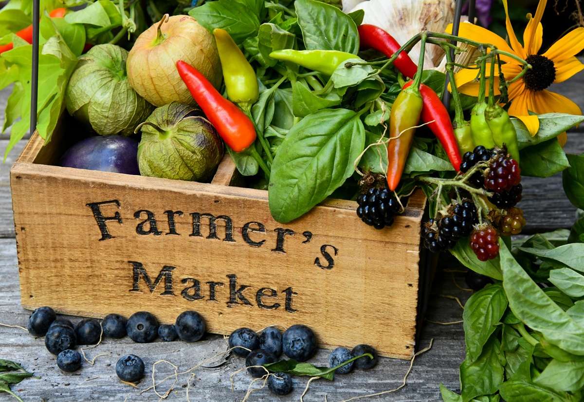 A photo of a box wooden box filled with vegetables and a sunflower. Farmer's Market is burned into the side. Blueberries are scattered in front of the box.